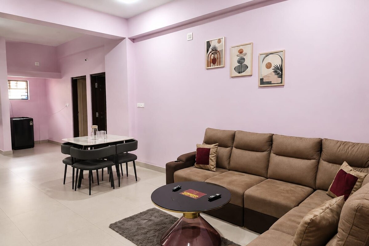 Una sala de estar con un sofá marrón, una mesa redonda de centro y una mesa de comedor con sillas. Hay tres obras de arte enmarcadas en la pared.