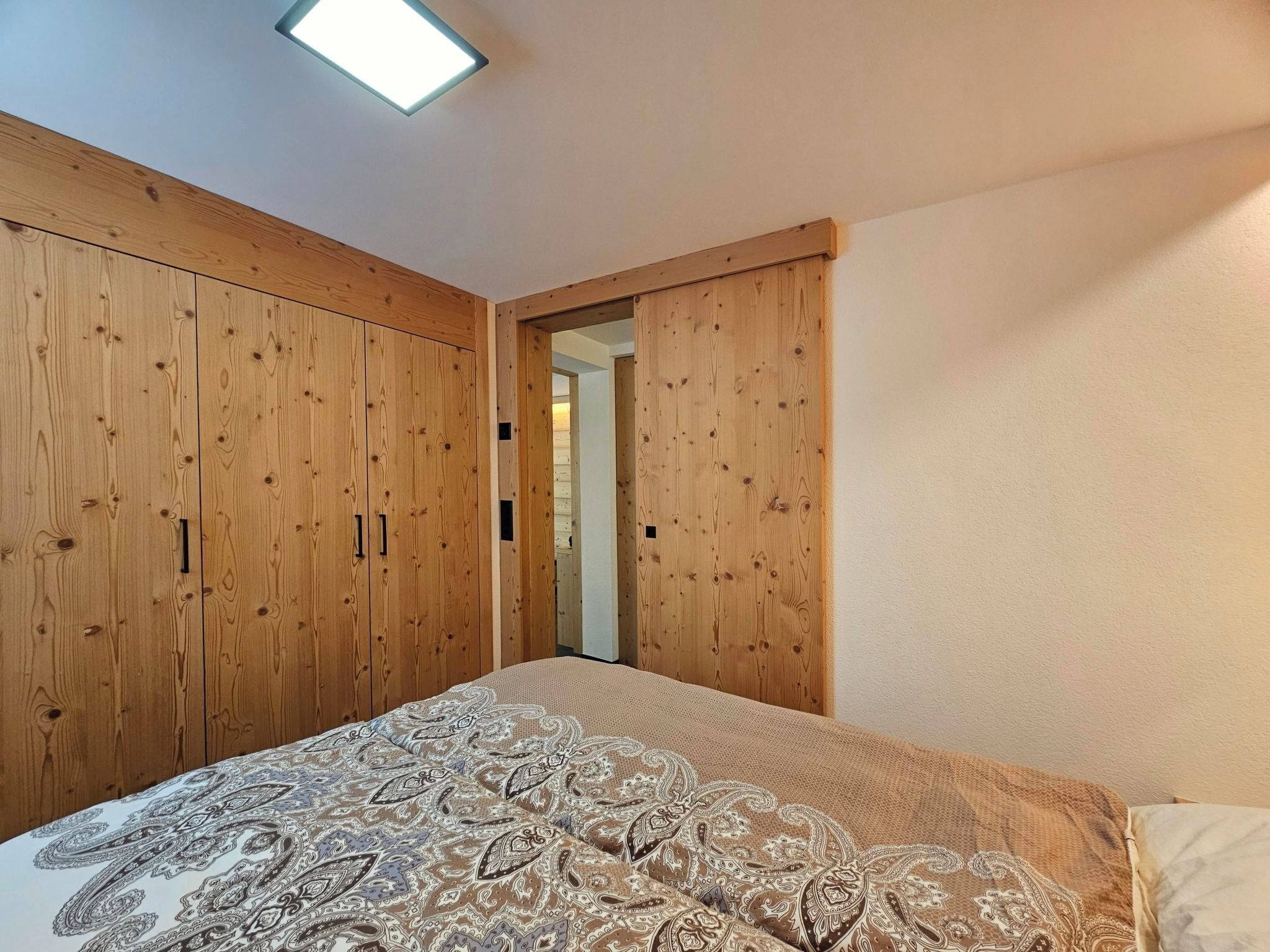 A bedroom with a bed, wooden wardrobe, and a door leading to another room.