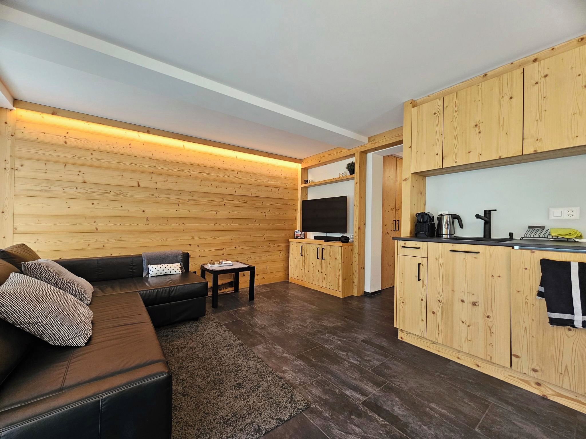 A modern living space with a wooden wall, a black sofa, a small table, and a kitchen area with wooden cabinets and a countertop.
