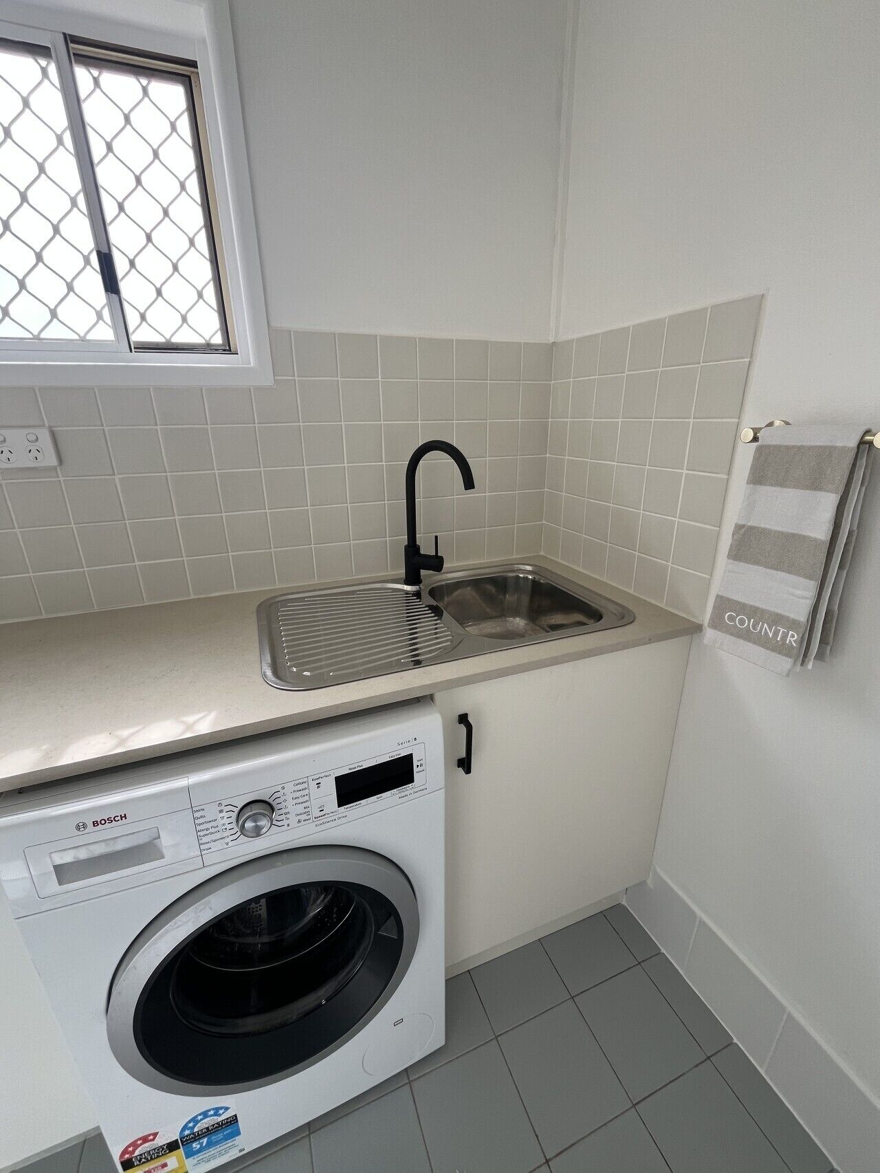 A compact kitchen with a Bosch washing machine, a sink, and a window with a metal screen.