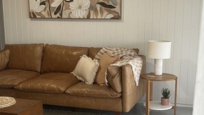 A living room with a brown leather sofa, a woven rug, a small wooden side table with a lamp, and a wall-mounted floral artwork. - Aelia in Peregian Beach (Peregian Beach)