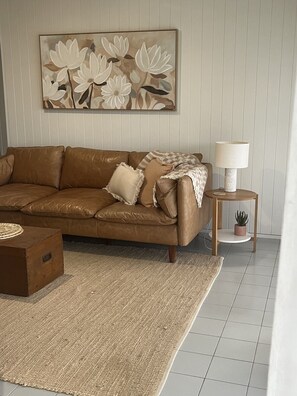 Ein Wohnzimmer mit braunem Ledersofa, einem gewebten Teppich, einem kleinen Holztisch mit Lampe und einem Wandbild mit floralem Muster.