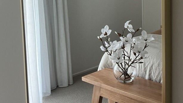 A bedroom with a bed, a wooden nightstand, a vase with flowers, a painting on the wall, and a window with curtains. - Aelia in Peregian Beach (Peregian Beach)
