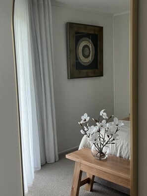 A bedroom with a bed, a wooden nightstand, a vase with flowers, a painting on the wall, and a window with curtains.