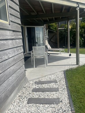 A covered patio area with a concrete floor, a white chair, a bench, and a small bird on the ground.