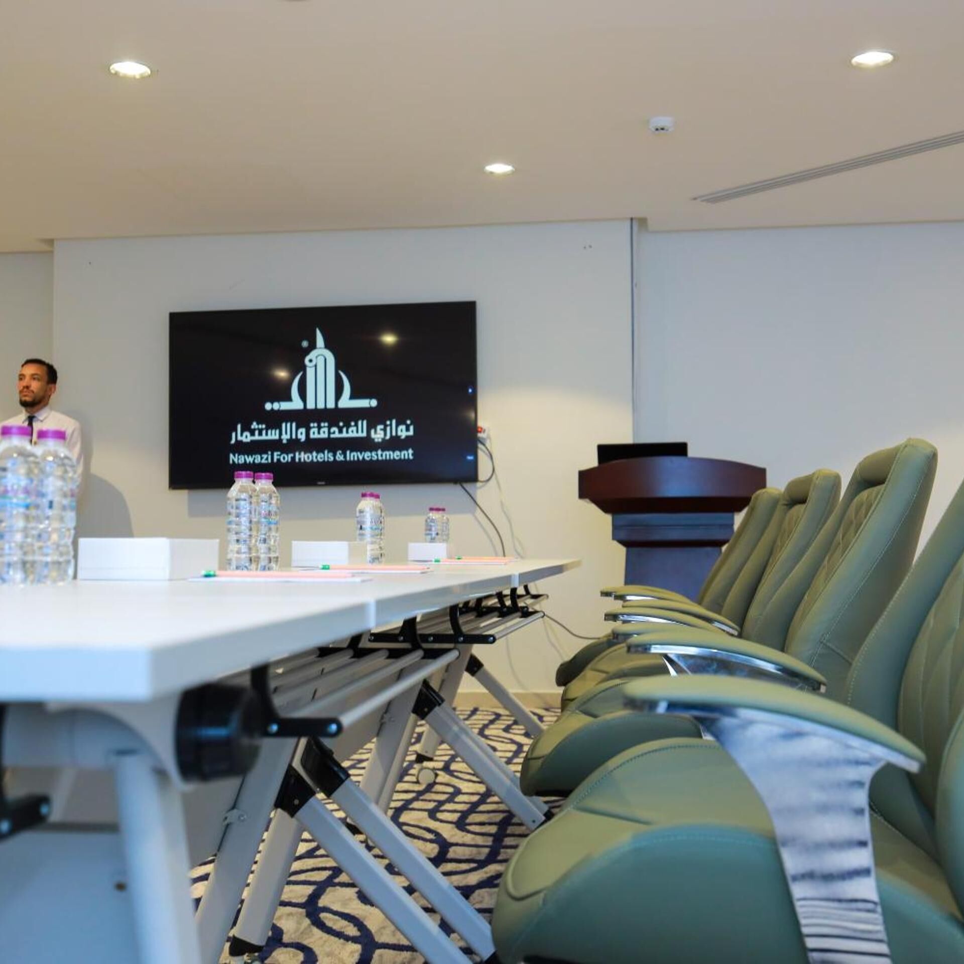 A conference room with a presentation screen, a table with water bottles, and green upholstered chairs.