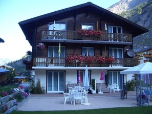A two-story wooden building with balconies, outdoor seating, and a mountain backdrop. - Apartment Anis by Interhome (Saas-Almagell)