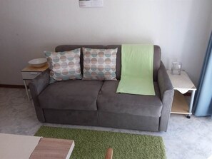 A grey sofa with patterned pillows and a green throw, a small white table with a bowl, and a green rug in a room with a white wall and a blue curtain. - Apartment Anis by Interhome (Saas-Almagell)