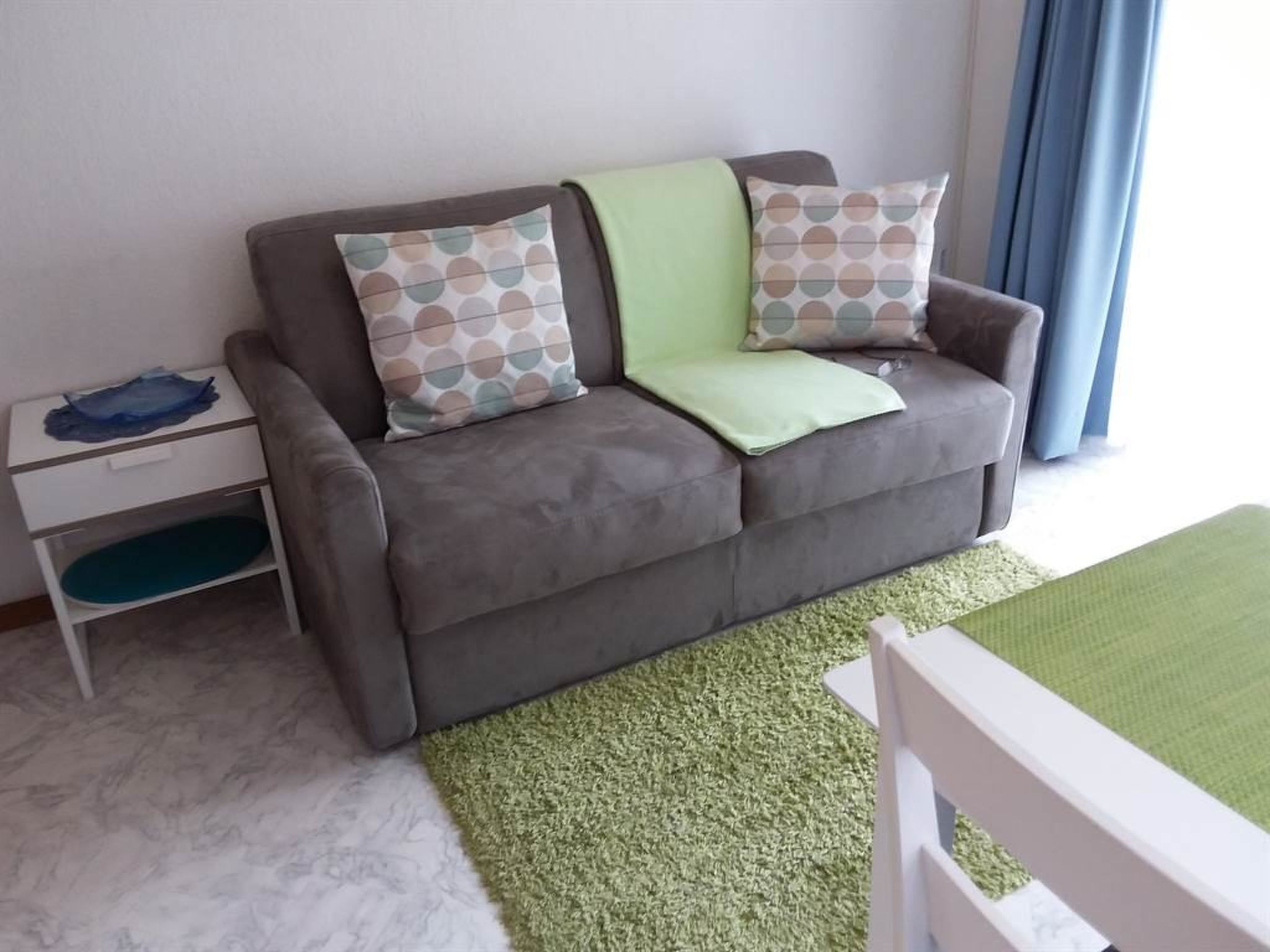 Un sofá gris con cojines estampados, una mesita auxiliar blanca y una alfombra verde.