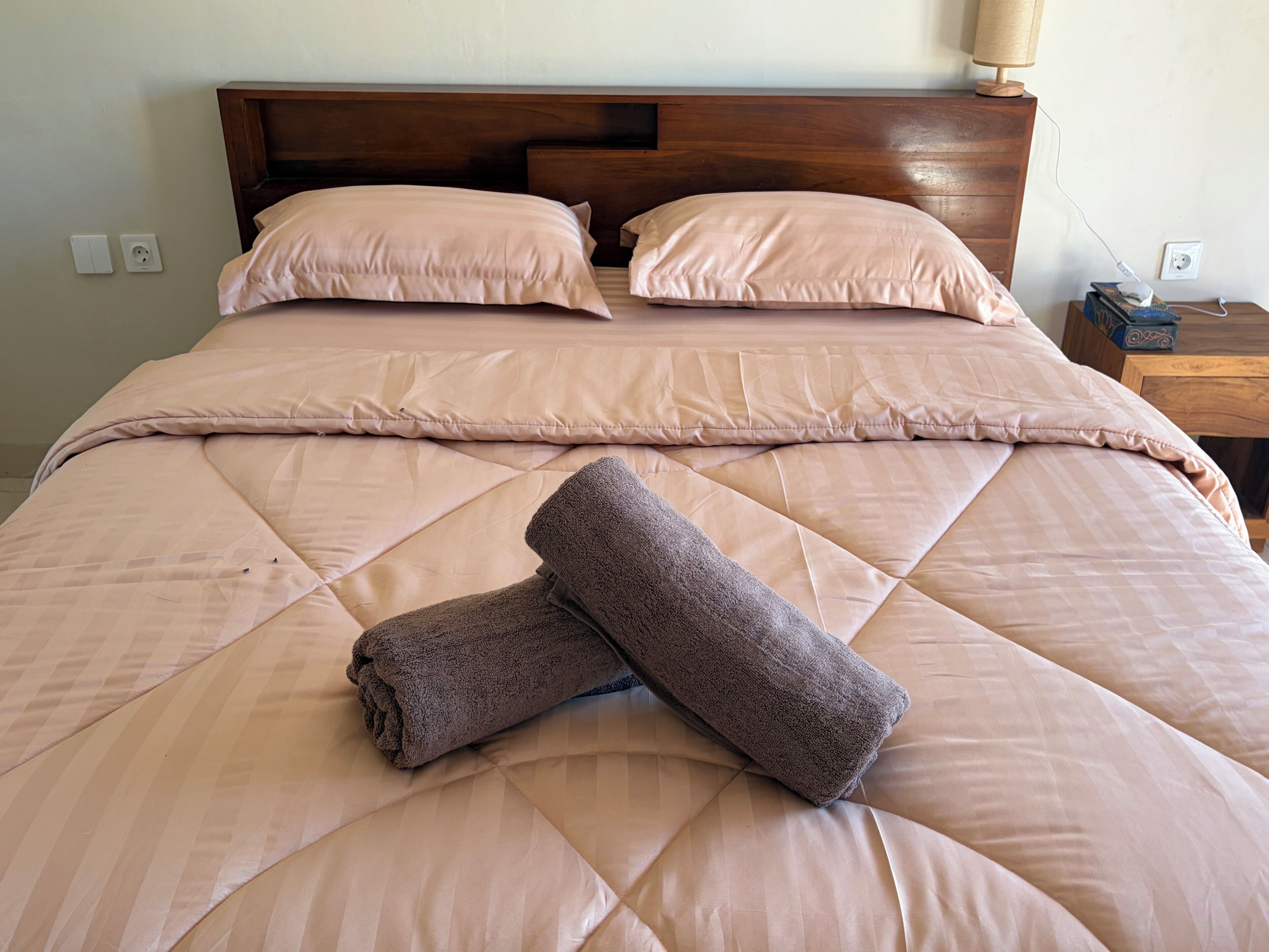 A neatly made bed with a quilted cover, two pillows, and two rolled towels.
