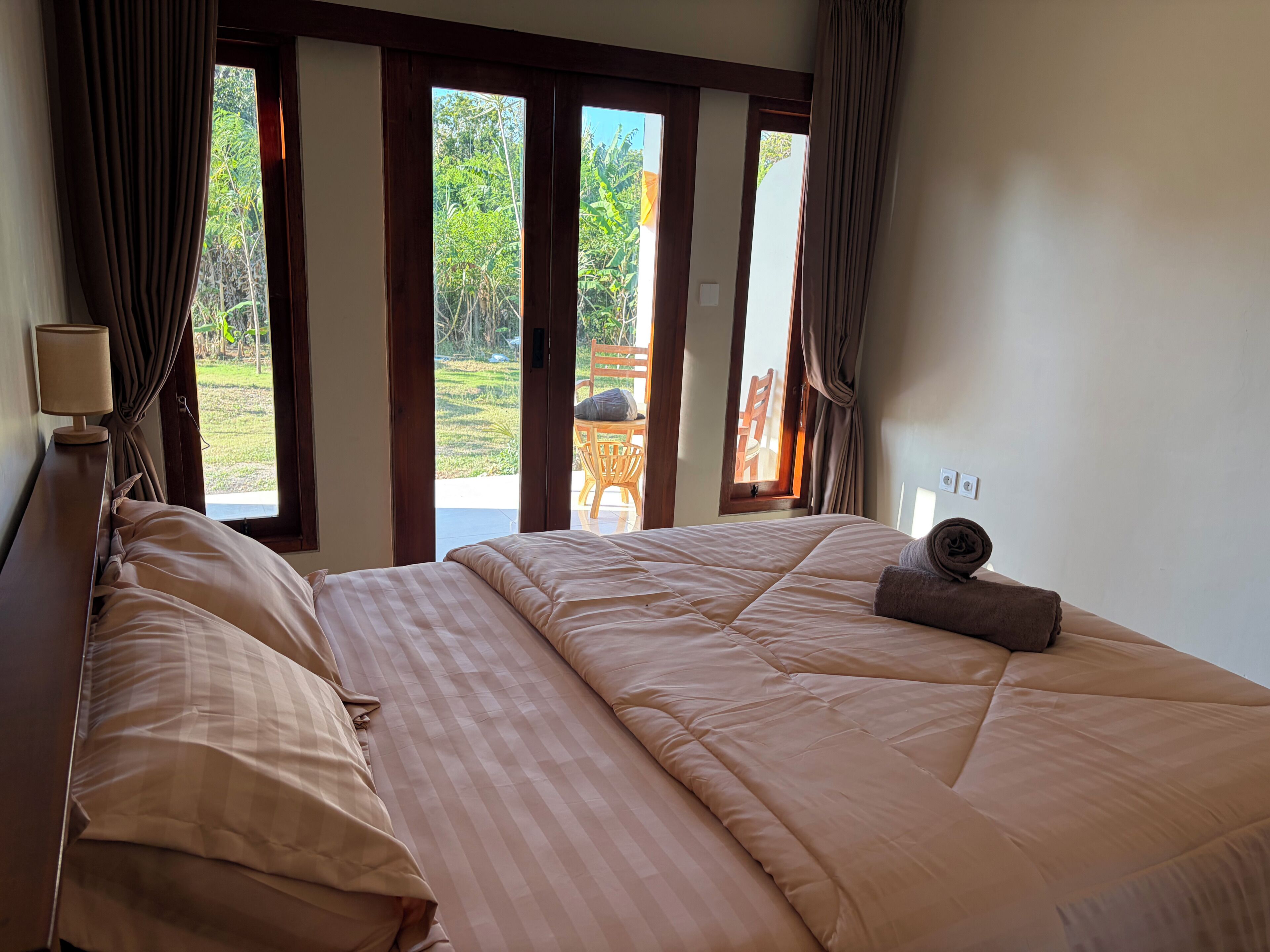 A bedroom with a bed, curtains, a lamp, and a view of the outdoors.