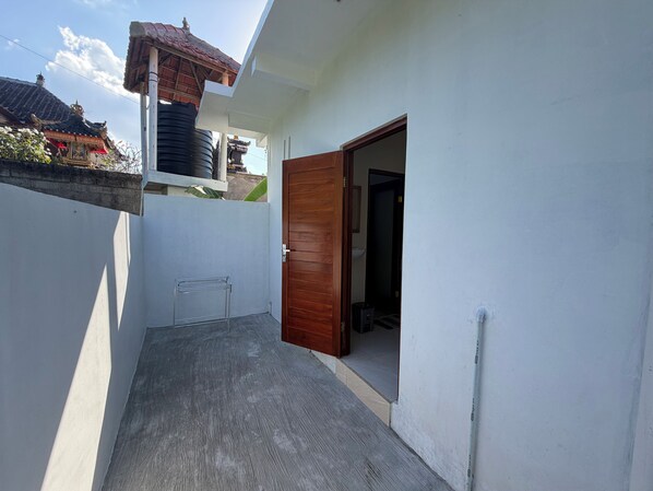 A narrow outdoor corridor with a wooden door leading to a dark interior. A metal rack is visible on the left, and a white wall runs along the right side.
