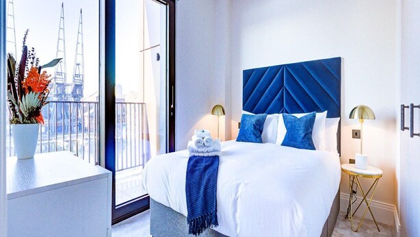 A hotel room with a large bed, a view of a cityscape, a balcony with plants, and a small table with a lamp. - Hybridresi Canary Wharf (London)