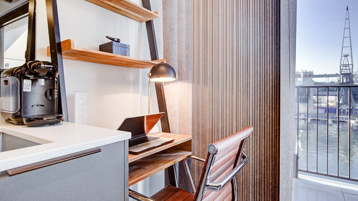 Une chambre d’hôtel moderne équipée d’un bureau, d’un ordinateur portable, d’une machine à café et offrant une vue sur la ville grâce à de grandes fenêtres.