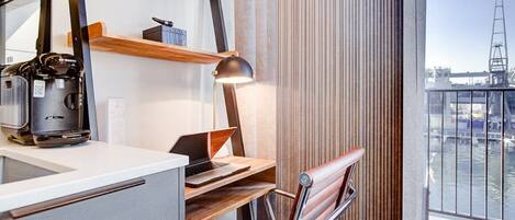 Une chambre d’hôtel moderne équipée d’un bureau, d’un ordinateur portable, d’une machine à café et offrant une vue sur la ville grâce à de grandes fenêtres.