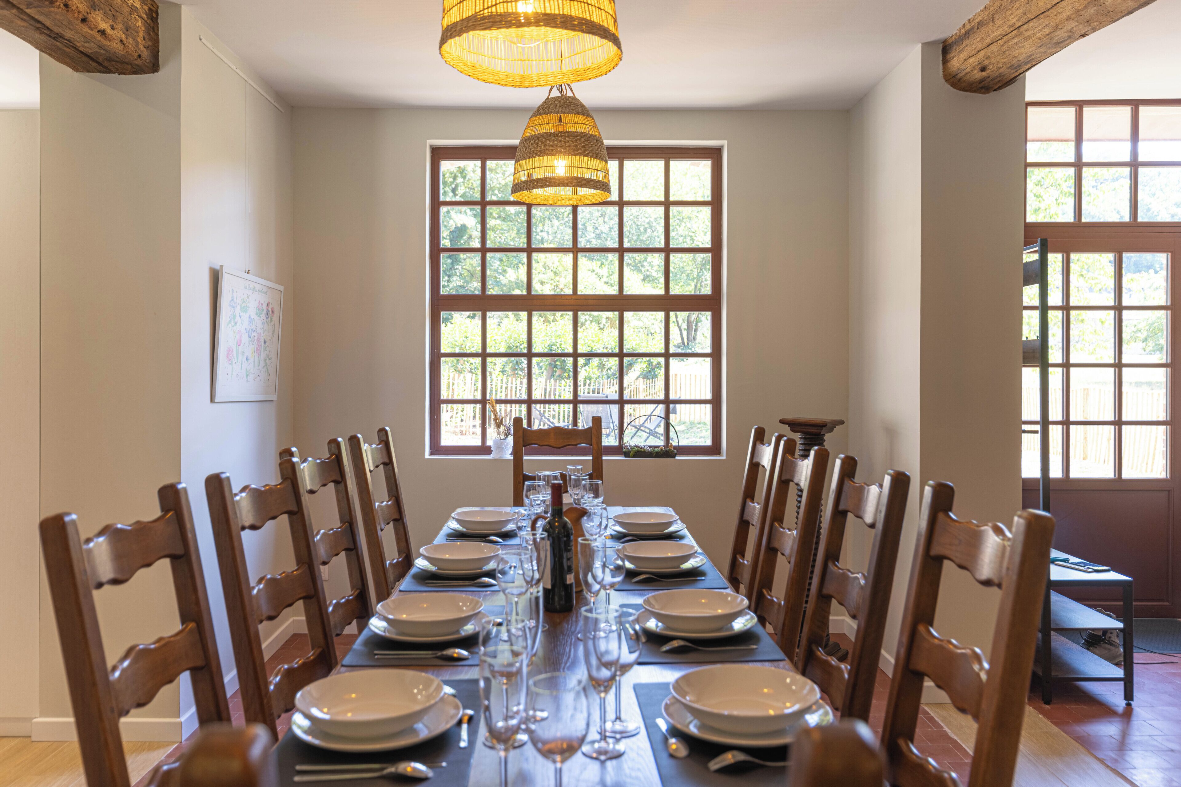 A dining room with a long table set for a meal, flanked by wooden chairs, a hanging light fixture, and a window with a view of greenery.