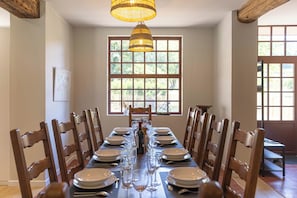 Une salle à manger avec une longue table dressée pour un repas, entourée de chaises en bois, un luminaire suspendu et une fenêtre donnant sur un paysage verdoyant.