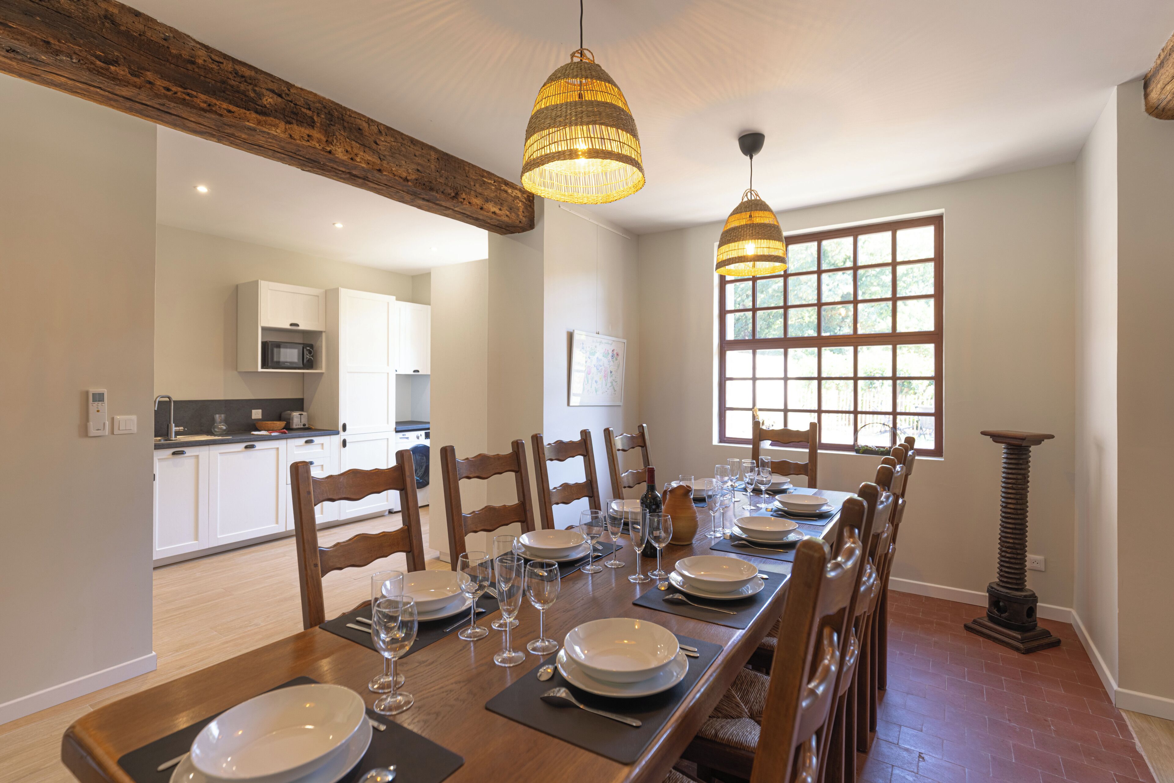 Une salle à manger avec une longue table dressée pour six personnes, des chaises en bois et deux lampes suspendues.