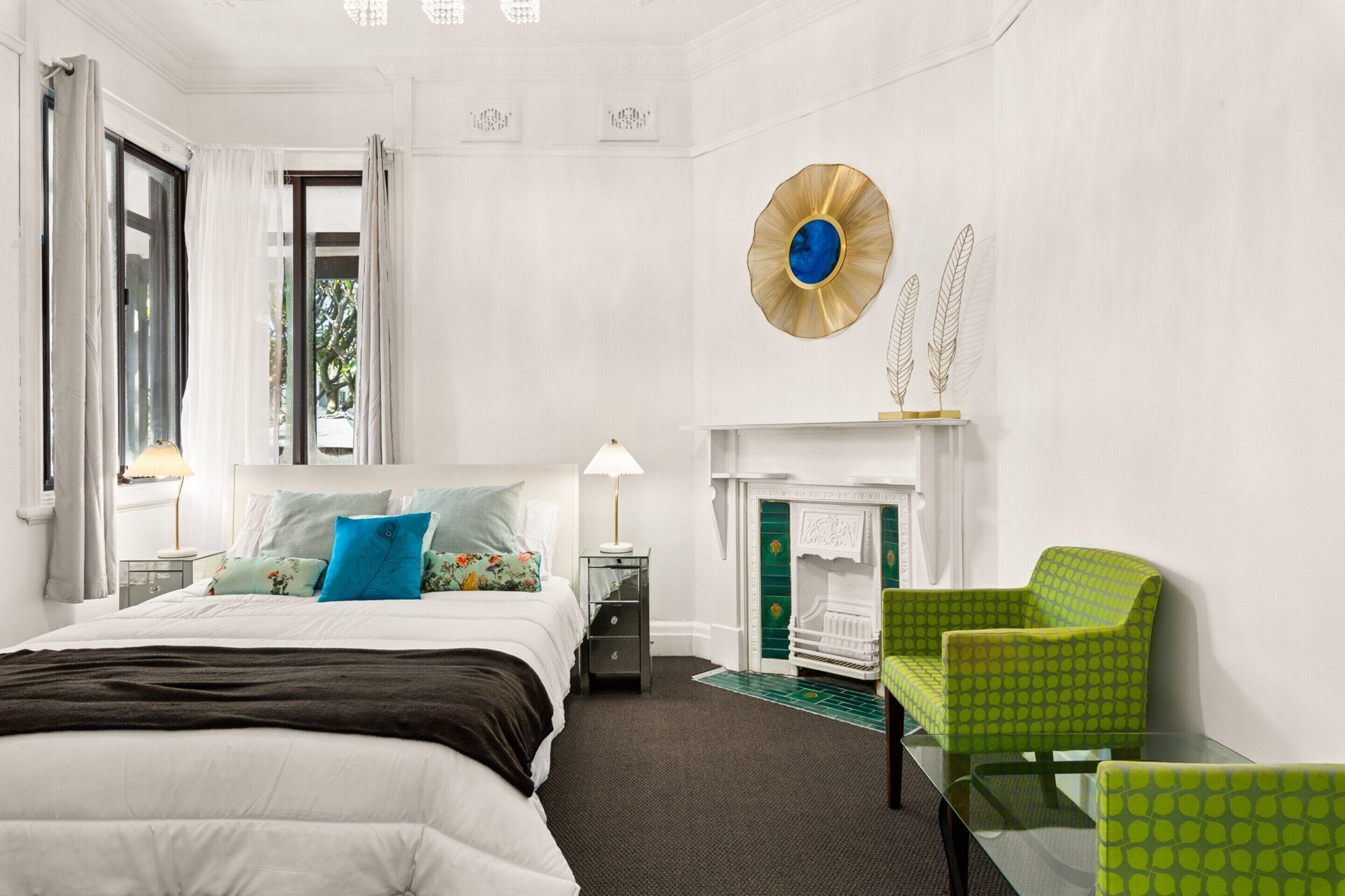 A hotel room with a bed, a nightstand, a lamp, a green armchair, a fireplace, and a decorative wall piece.