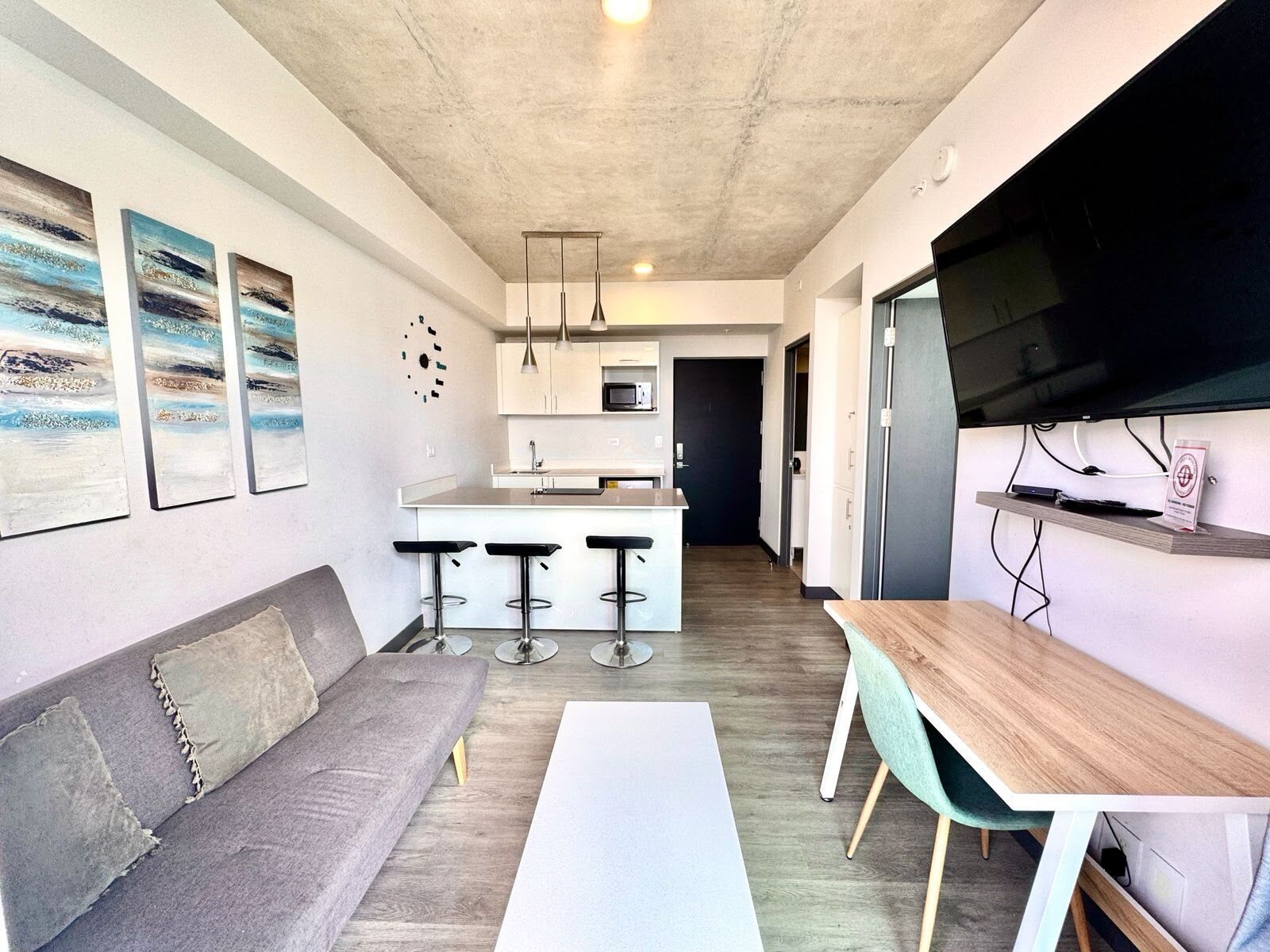 A modern kitchen with white cabinets, a central island with stools, a flat-screen TV mounted on the wall, and a grey sofa with a white coffee table.