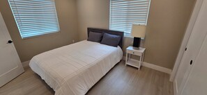 Un dormitorio con cama, mesita de noche, lámpara y dos ventanas con persianas.