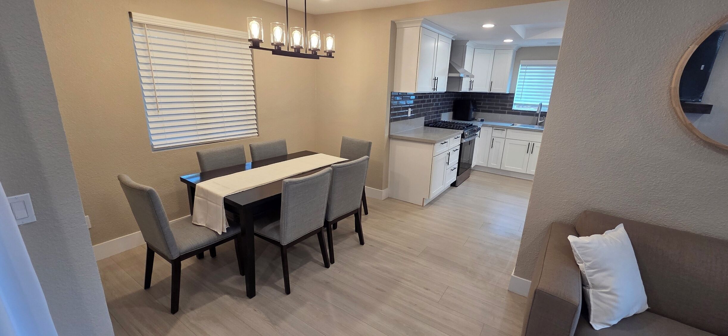 A dining area with a table and chairs, a kitchen in the background, and a view of the living room.