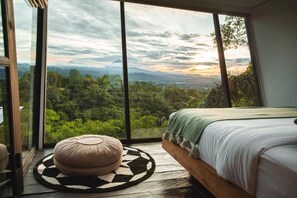 A bedroom with a large window, a bed, and a round ottoman.