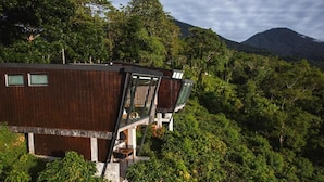 Um edifício moderno de madeira com varanda, rodeado por vegetação exuberante e uma montanha ao fundo.