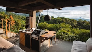 Uma área de cozinha ao ar livre com churrasqueira, pia e espaço para refeições, com vista para uma paisagem verdejante.