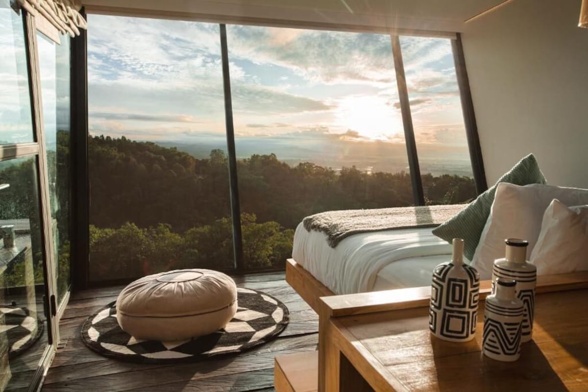 Un dormitorio con una ventana grande, una cama, un reposapiés redondo y jarrones decorativos.