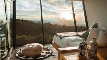 A bedroom with a large window, a bed, a round ottoman, and decorative vases.