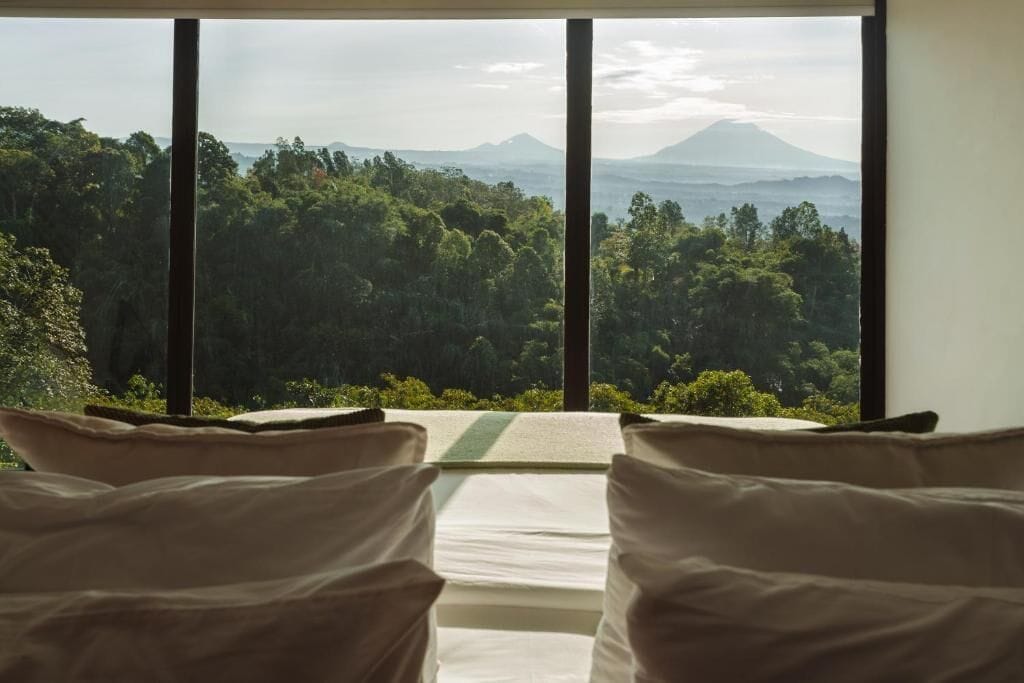 A room with a bed facing a scenic view of a forest and mountains.