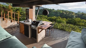 Cozinha externa com churrasqueira, pia e área de refeições, com vista para uma paisagem arborizada e exuberante.