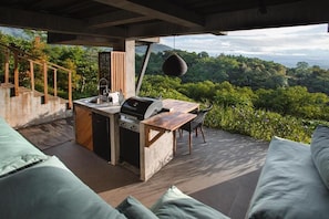 Un área de cocina al aire libre con una parrilla, un lavamanos y un comedor, con vistas a un paisaje boscoso exuberante.