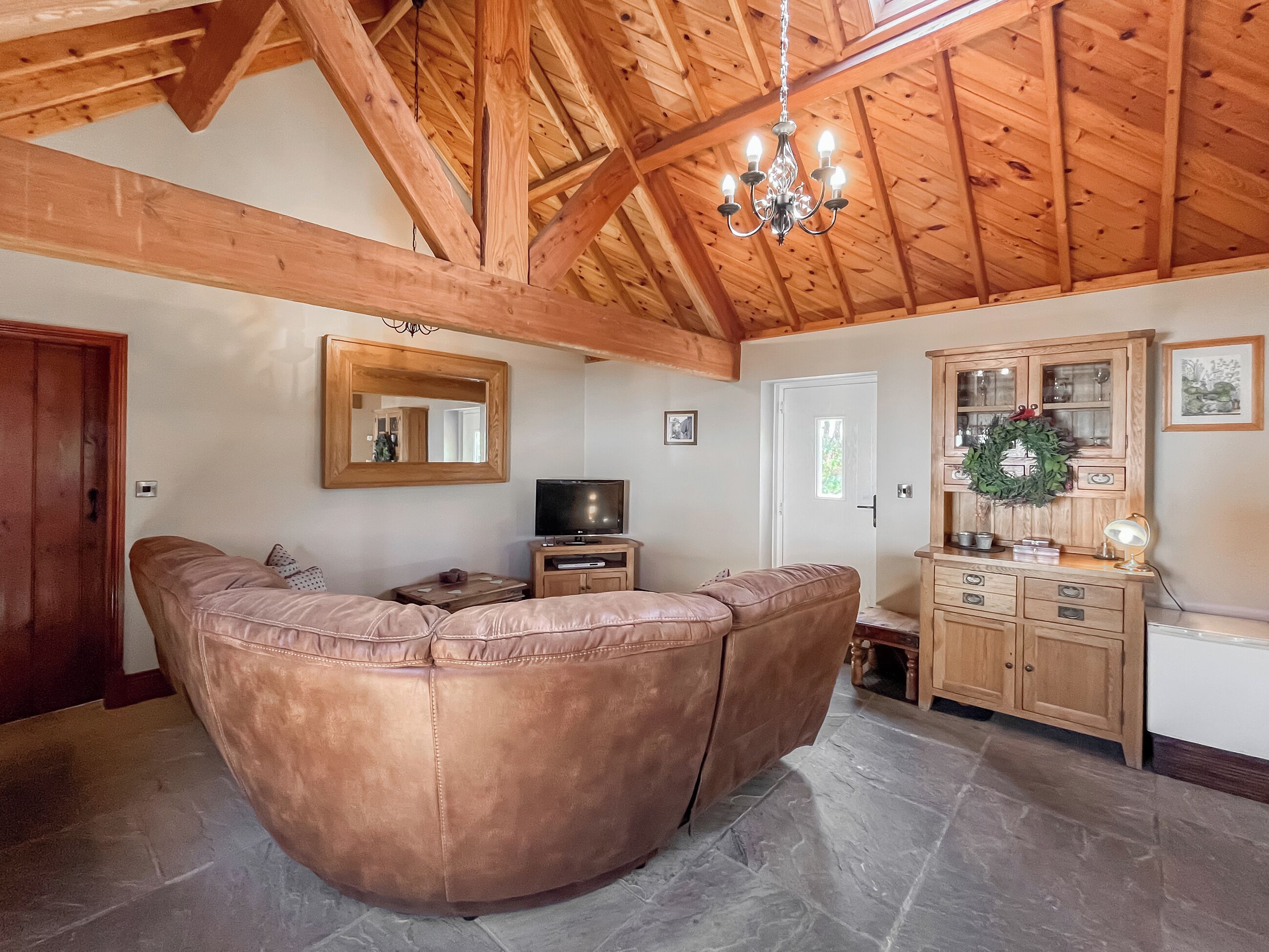 A living room with a curved leather sofa, wooden ceiling beams, a chandelier, and a wooden cabinet with glass doors.