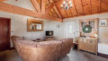 A living room with a curved leather sofa, wooden ceiling beams, a chandelier, and a wooden cabinet with glass doors.