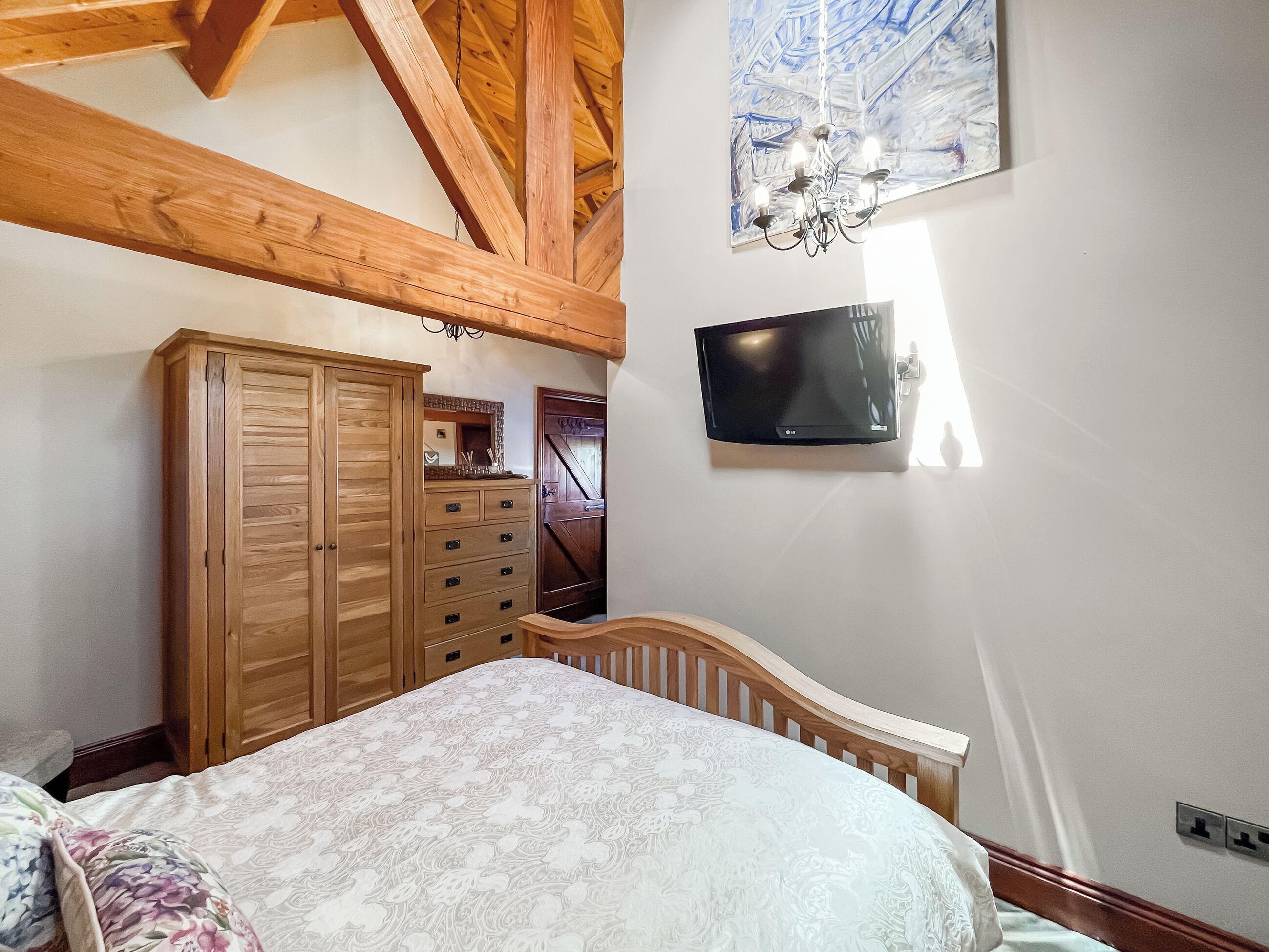 Une chambre avec une armoire en bois, un lit recouvert d’une couvre-lit à motifs floraux, un téléviseur fixé au mur et un lustre.