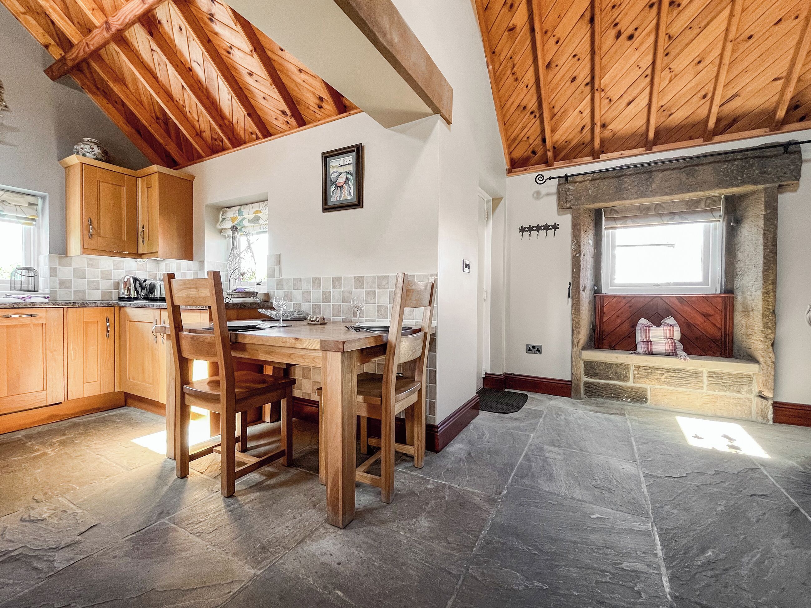 Une cuisine avec des meubles de rangement en bois, une table à manger avec des chaises, un sol en pierre et une cheminée avec un banc.