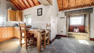 Une cuisine avec des meubles de rangement en bois, une table à manger avec des chaises, un sol en pierre et une cheminée avec un banc.