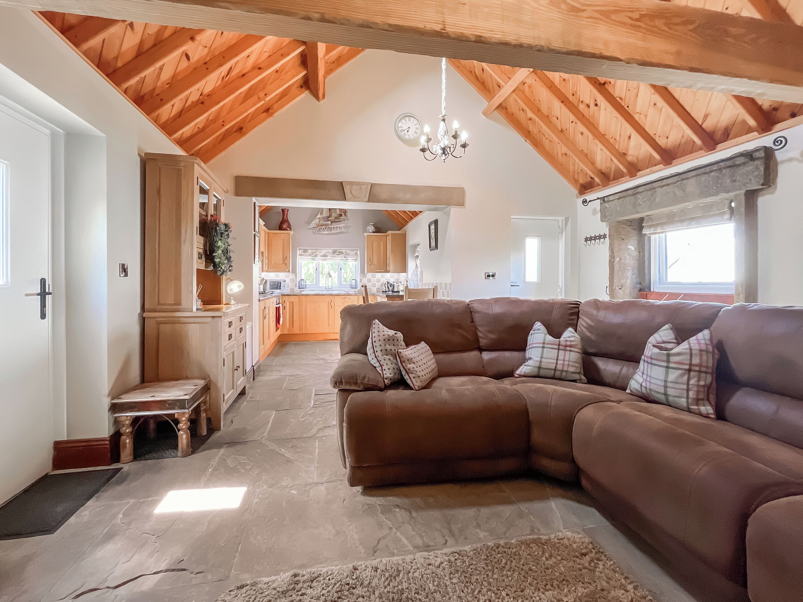 A spacious living area with a brown leather sofa, a wooden ceiling, and a view into the kitchen.