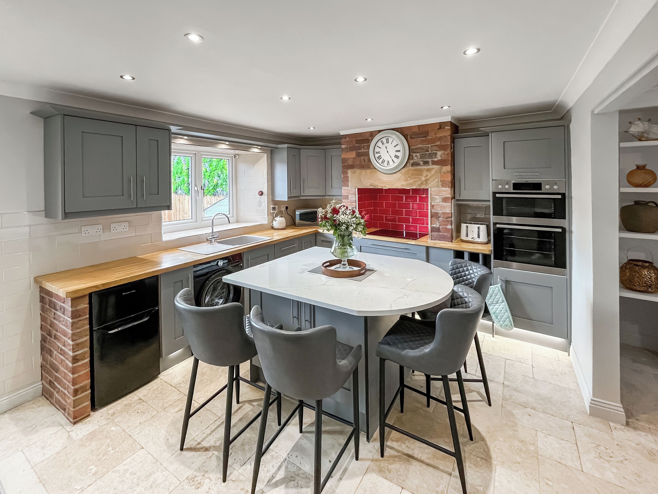 A modern kitchen with grey cabinetry, a central island with bar stools, a red brick backsplash, and built-in appliances.