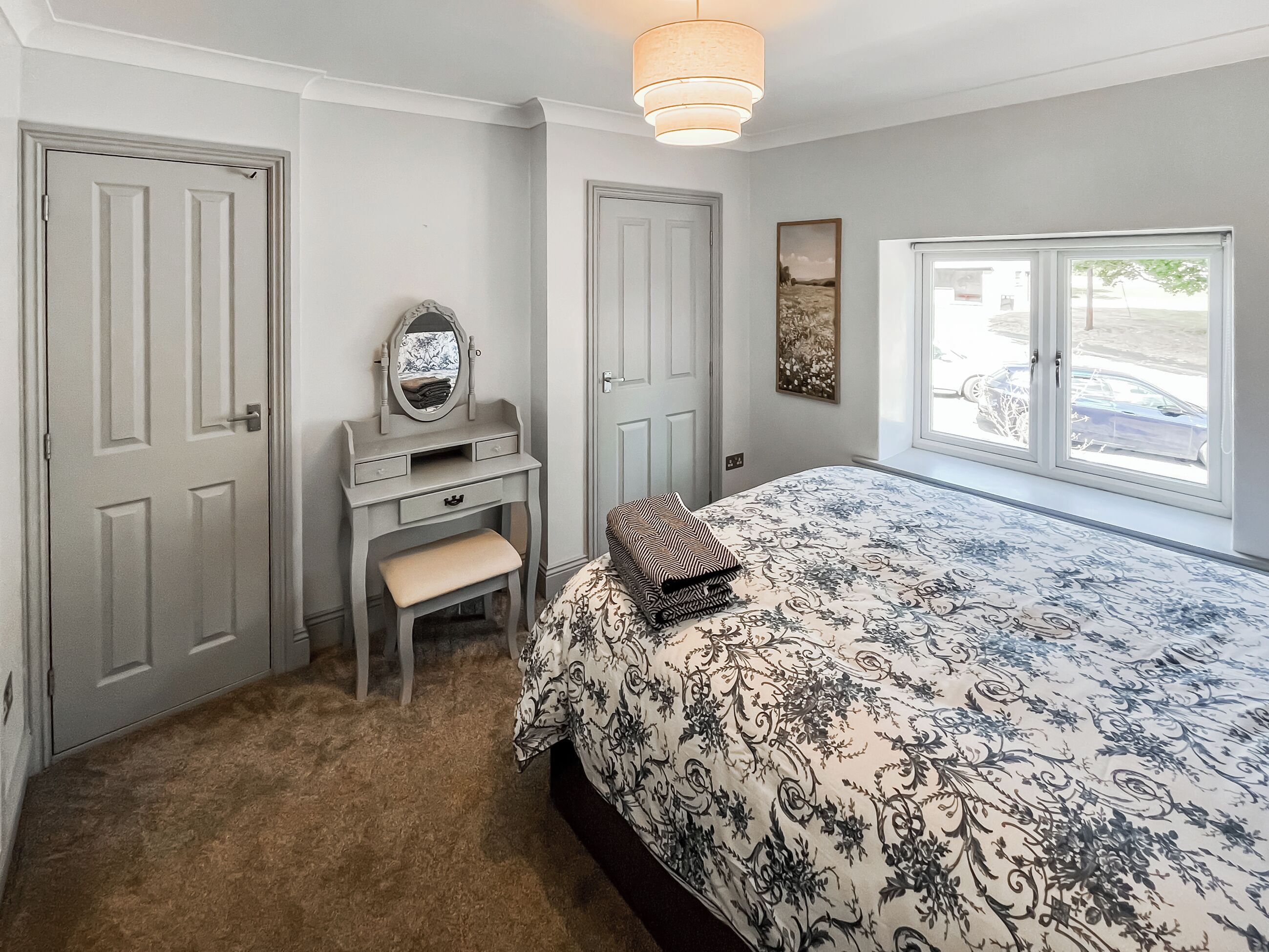 A bedroom with a bed, a vanity with a mirror, a bench, and a window with a view of the outside.