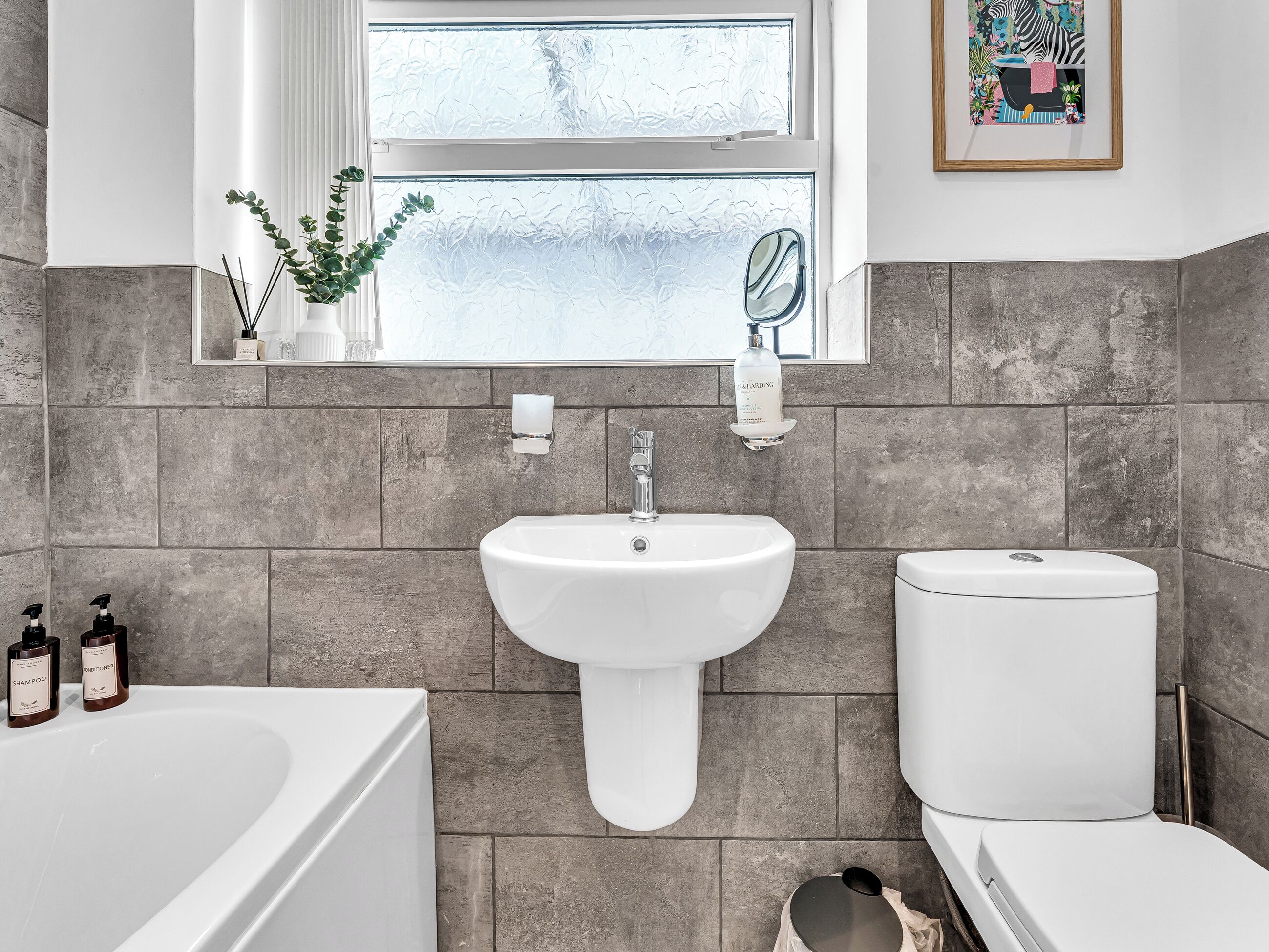 A bathroom with a white sink, a toilet, and a bathtub. There is a window with frosted glass, a mirror, and a framed picture on the wall.