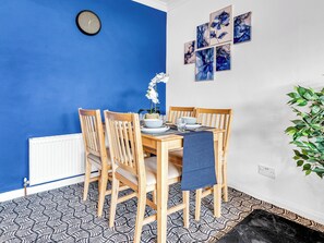 Uma sala de jantar com parede azul, cadeiras de madeira, mesa posta com pratos, tigelas e copos, e um relógio na parede.