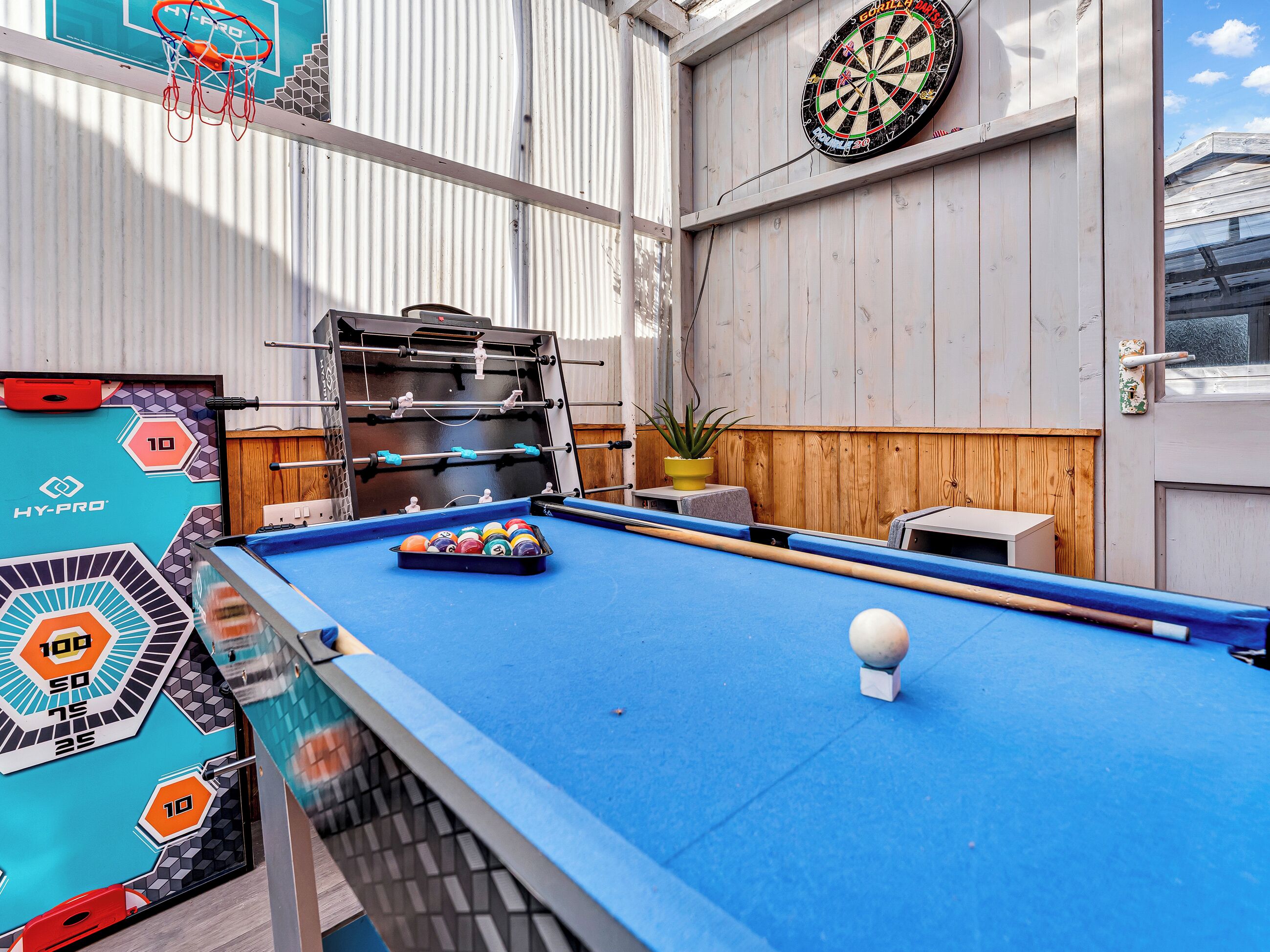 A pool table with a dartboard and a foosball table in a room with wooden walls and a window.