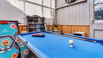 A pool table with a dartboard and a foosball table in a room with wooden walls and a window.