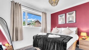 Une chambre à coucher avec un lit, une table de chevet, une lampe, une fenêtre avec des rideaux et des œuvres d’art accrochées au mur.