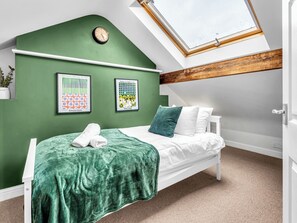 Ein Schlafzimmer mit schrägen Wänden, einem Oberlicht, einem Bett mit grüner Decke, zwei Kissen und zwei gerahmten Kunstwerken an der Wand.
