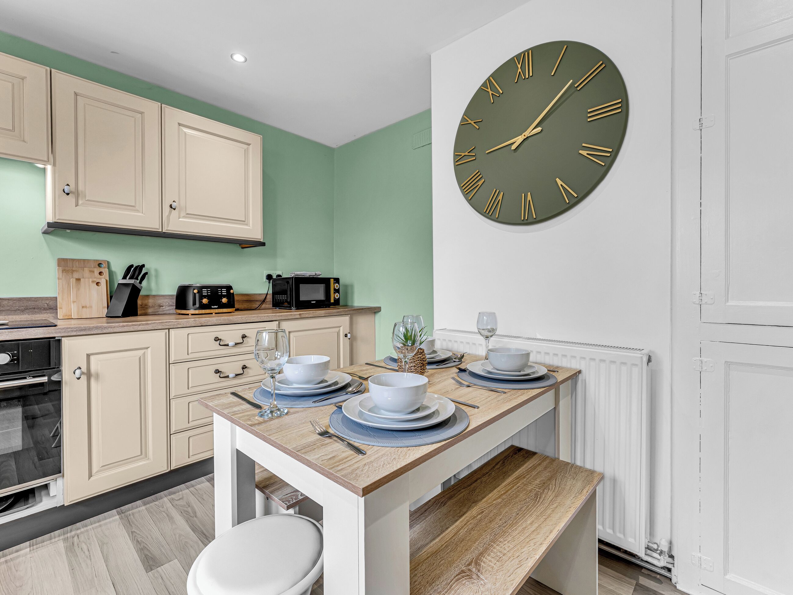 A kitchen with a dining table set for two, a microwave, and a clock on the wall.