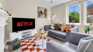 A living room with a television displaying 'NETFLIX,' a sofa, a coffee table, and a view of greenery outside. - 3 bedroom accommodation in Leek (Leek)